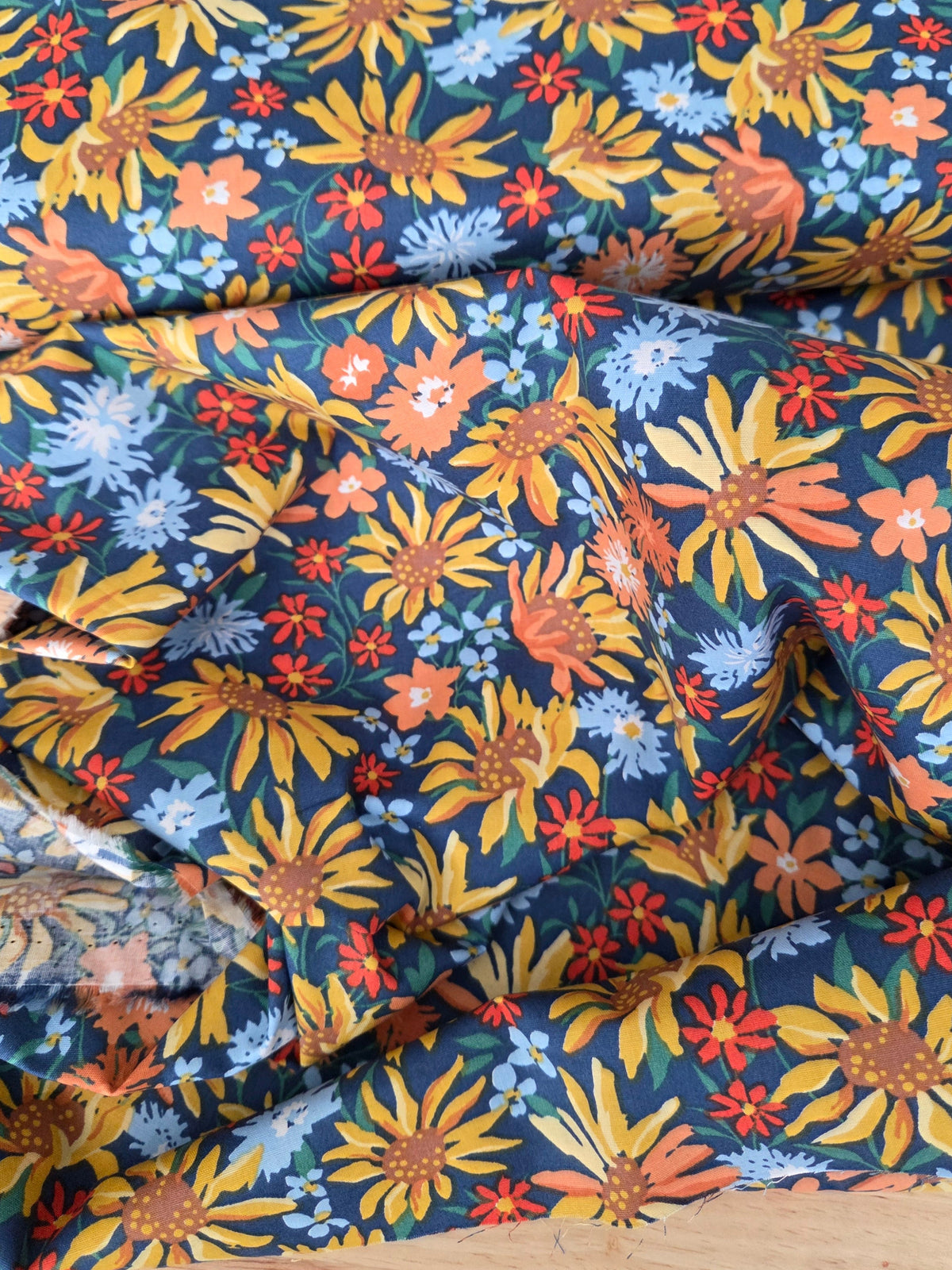 A close-up of draped fabric with a dense floral print on a dark teal-blue background, featuring yellow sunflowers, orange flowers, and light blue blossoms, laid over a light wood surface.