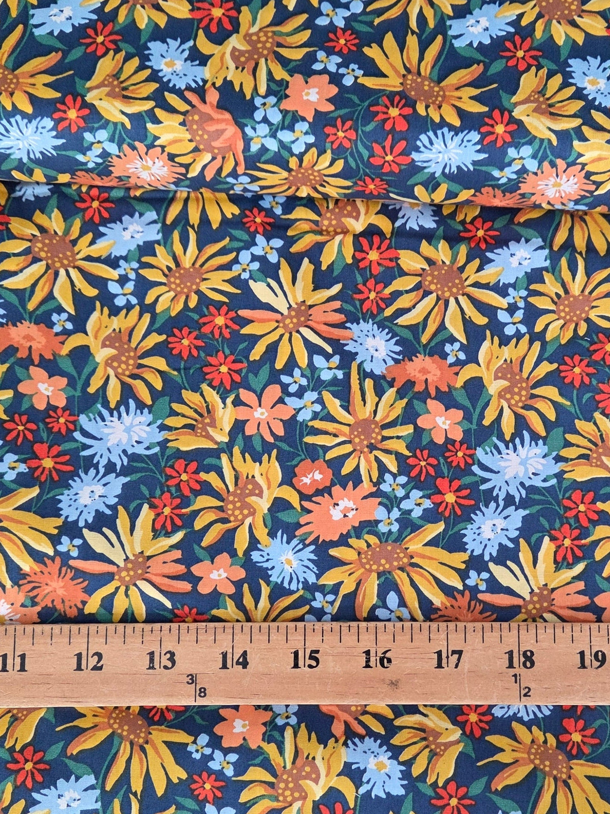 A close-up of a floral fabric with a repeating pattern of yellow, orange, and light blue flowers on a dark teal background, with a wooden ruler laid horizontally to show scale.