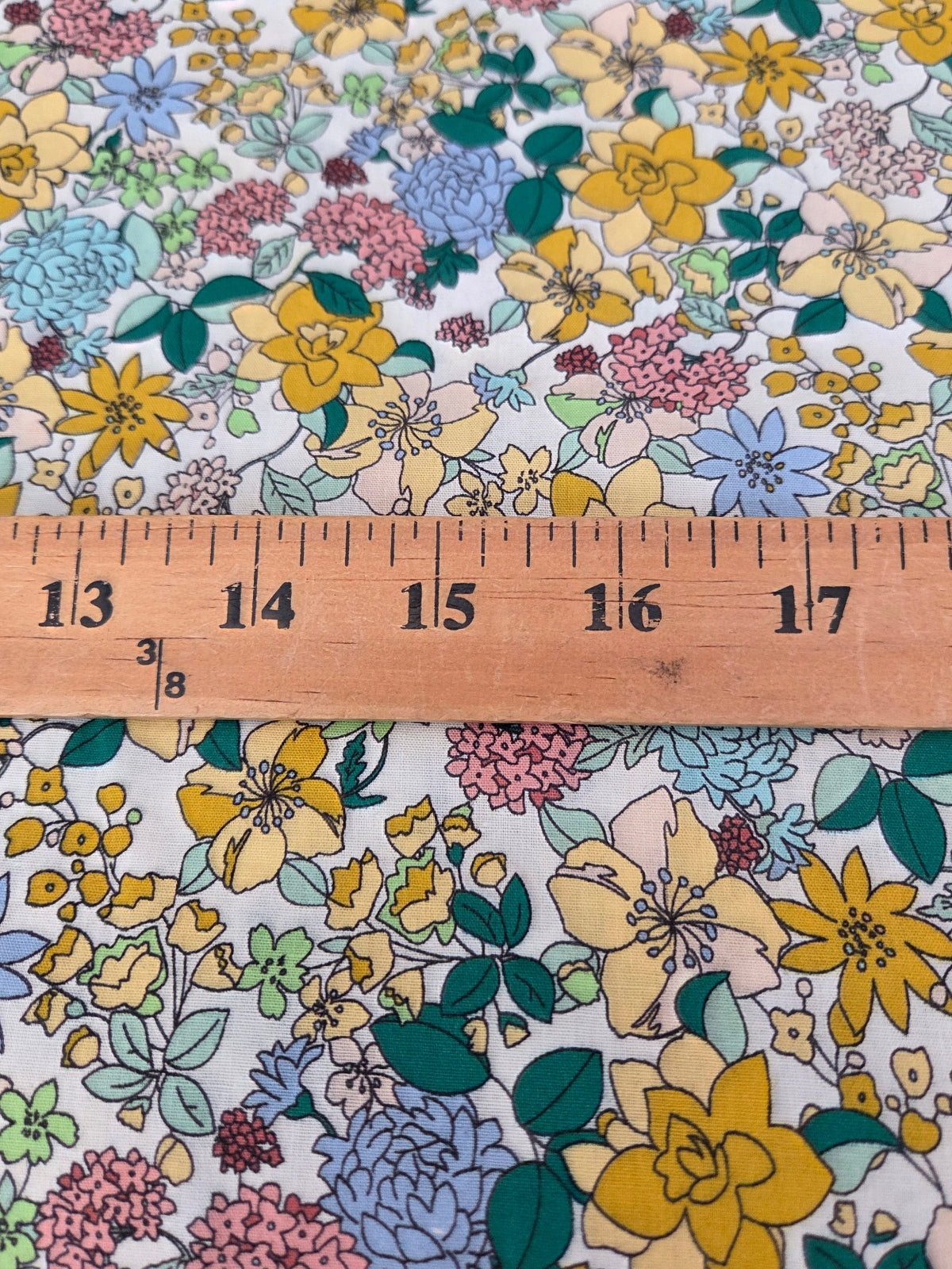 A close-up of a floral fabric with yellow, blue, pink, and green flowers on a white background, with a wooden ruler laid horizontally to show scale.