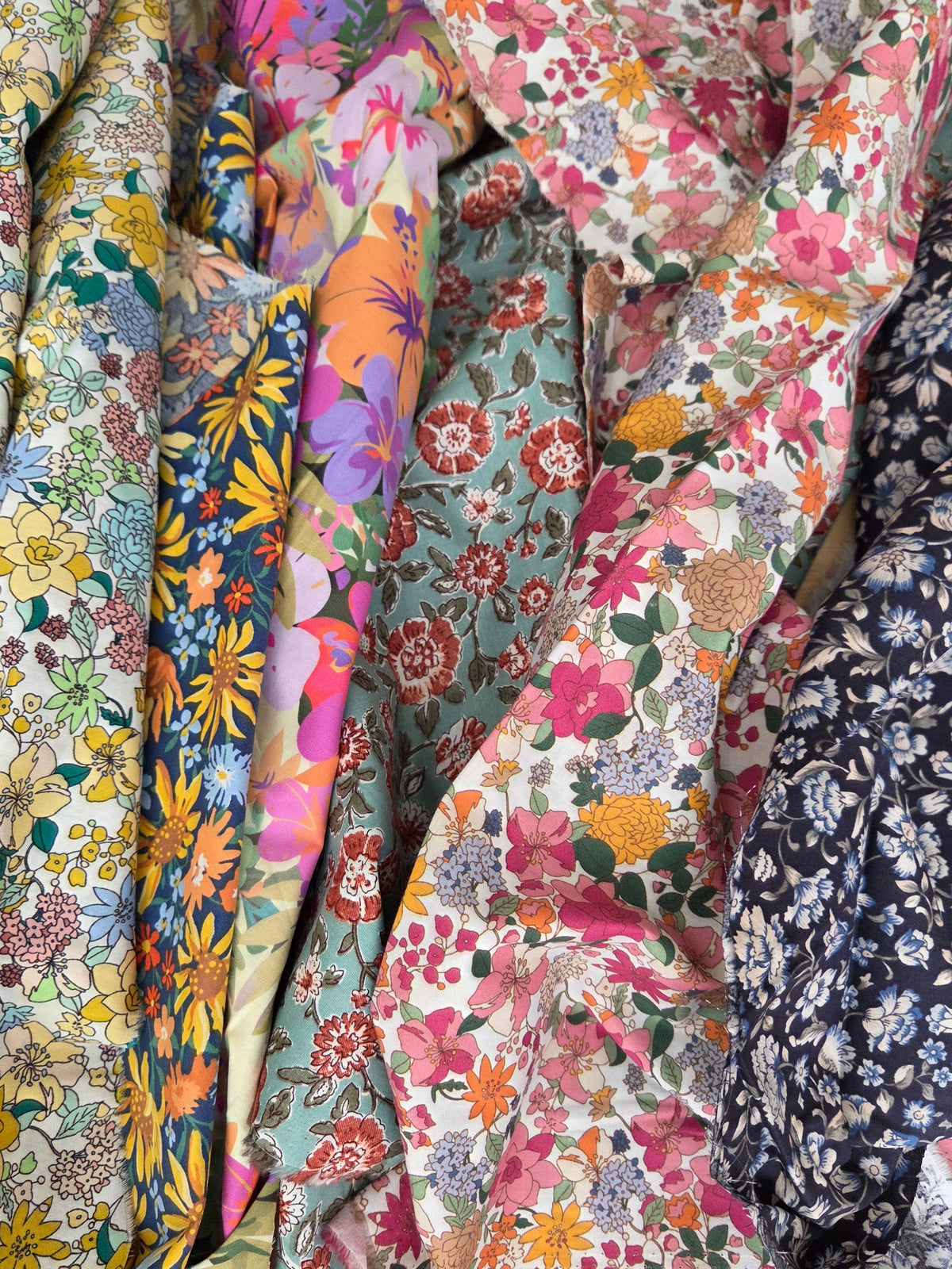 A close-up of several folded and draped floral fabrics in different patterns and colors, including pink, yellow, blue, green, and navy on white backgrounds.
