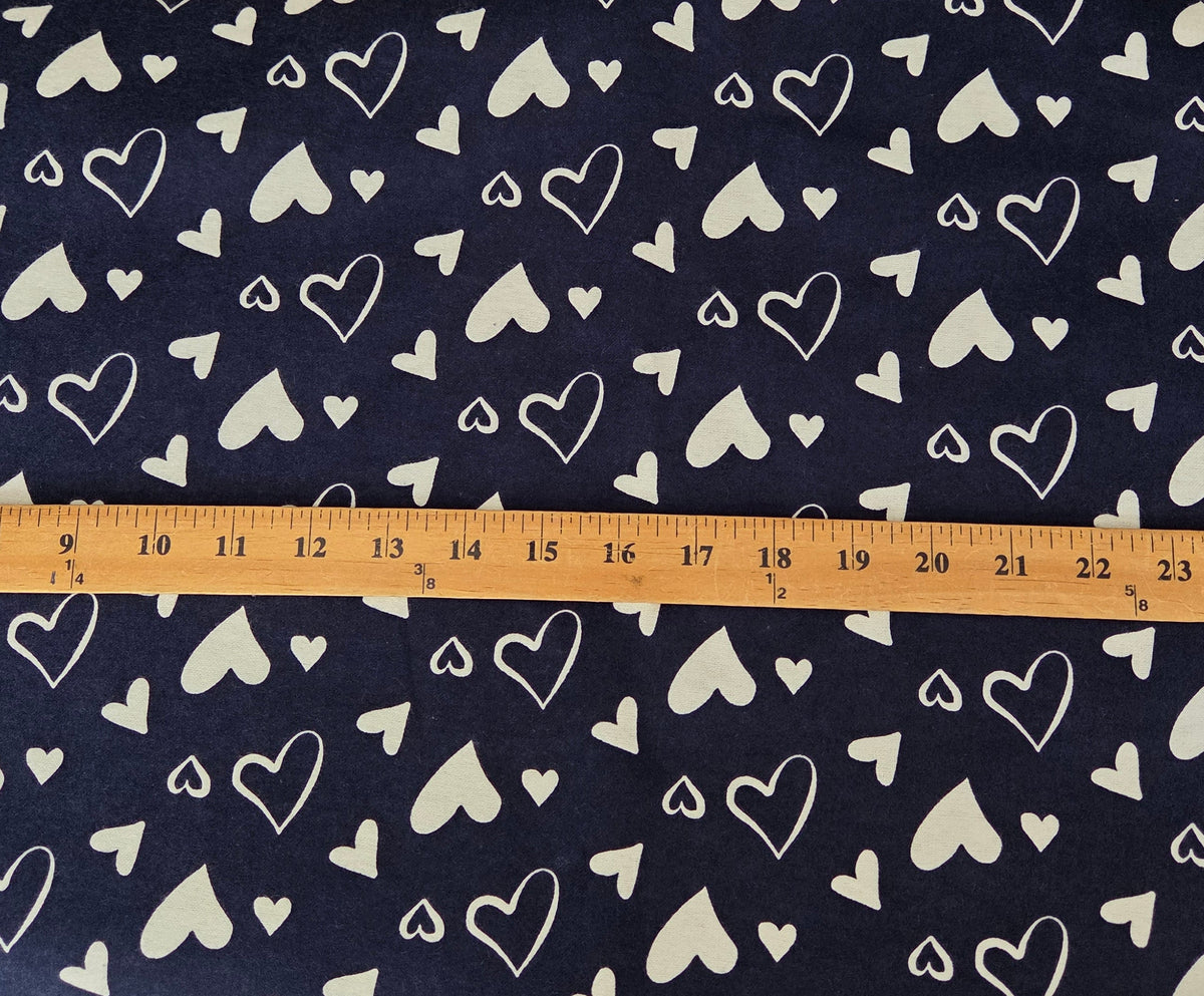 A navy blue fabric with a white heart pattern is laid flat on a wooden table, with a wooden ruler placed horizontally across the center to show the scale of the print. Other household items are visible in the background.