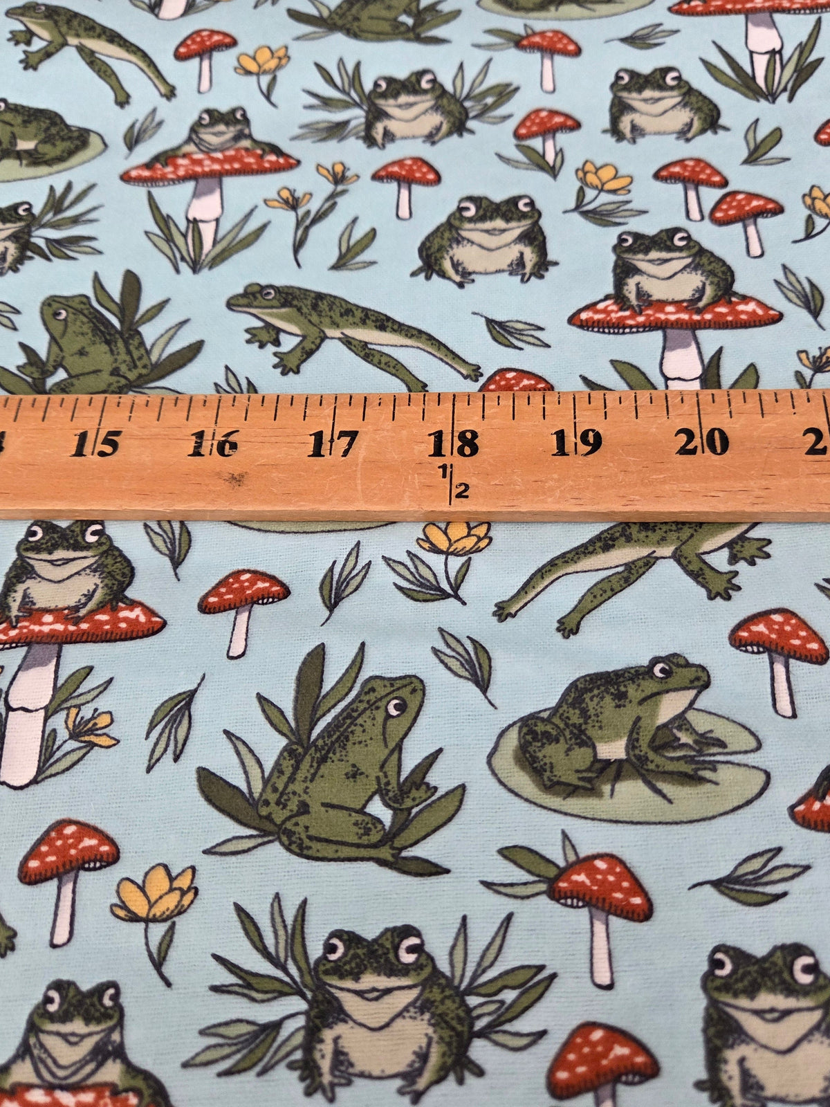 Light blue fabric with a repeating pattern of green frogs, red-and-white mushrooms, and small yellow flowers, shown with a wooden ruler across the middle for scale. LA FInch Fabrics. flannel by the yard, US fabric shop