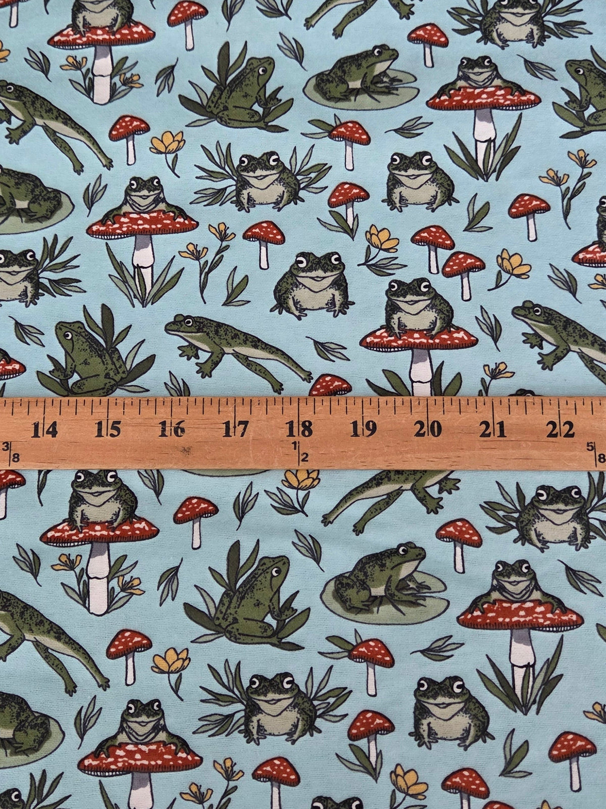 A light blue fabric with a repeating pattern of frogs,  and red-and-white mushrooms, shown with a wooden ruler across the middle to indicate scale cotton flannel by the yard