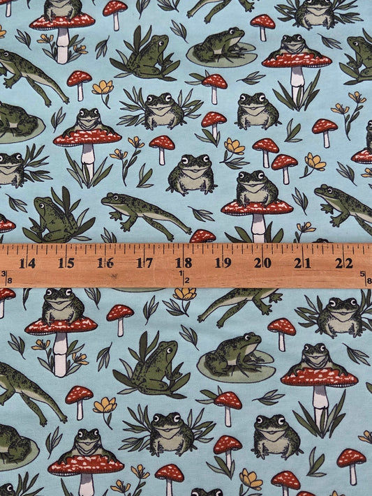 A light blue fabric with a repeating pattern of frogs,  and red-and-white mushrooms, shown with a wooden ruler across the middle to indicate scale cotton flannel by the yard