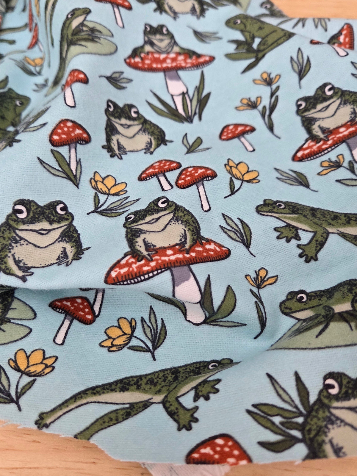 Light blue fabric with a whimsical frog and mushroom print, draped on a wooden surface. Fabric by the yard value line of flannels