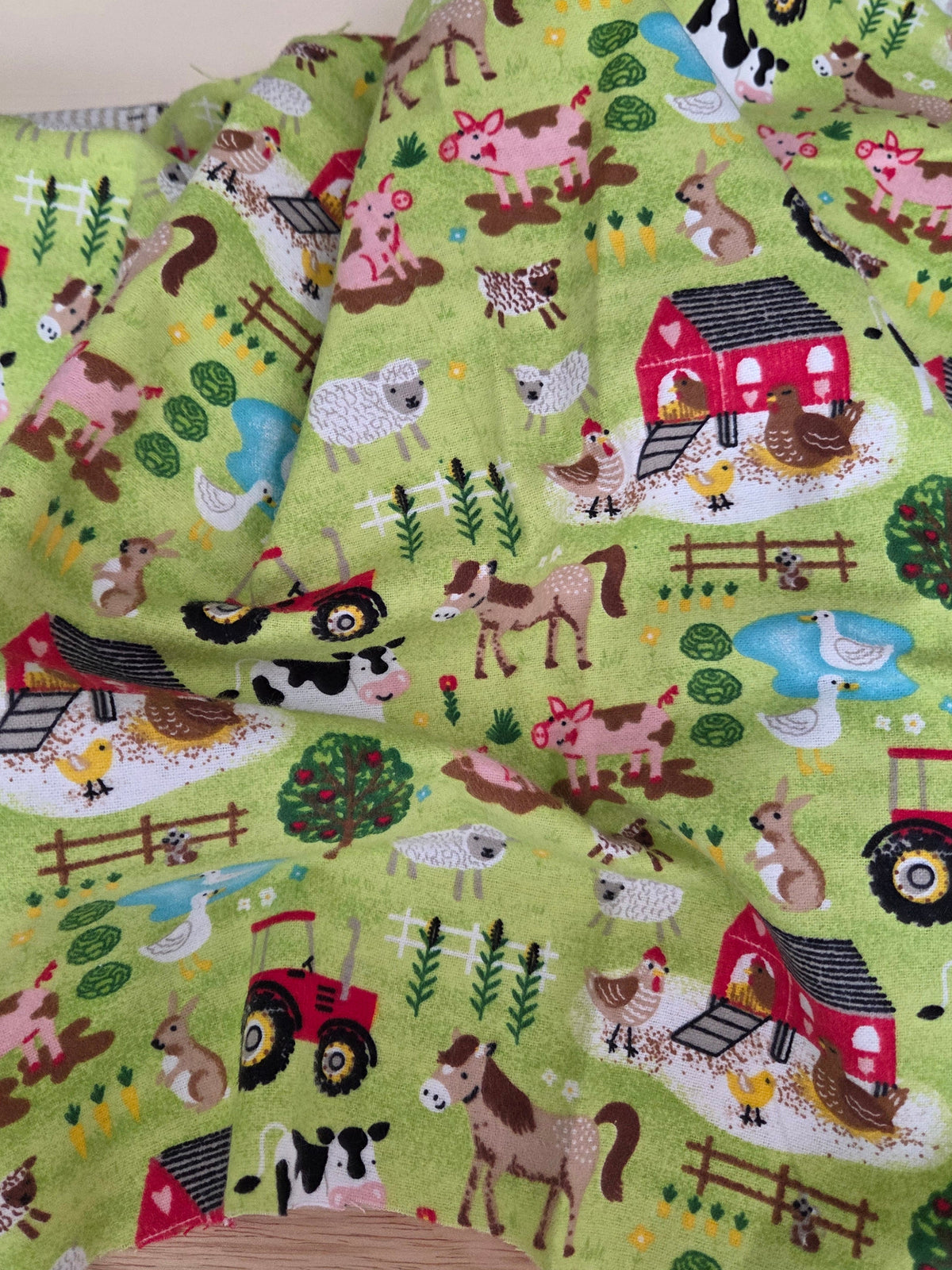 A folded piece of light green fabric with a colorful farm animal and vehicle print, displayed on a wooden surface next to a wooden ruler. LA FInch Fabrics