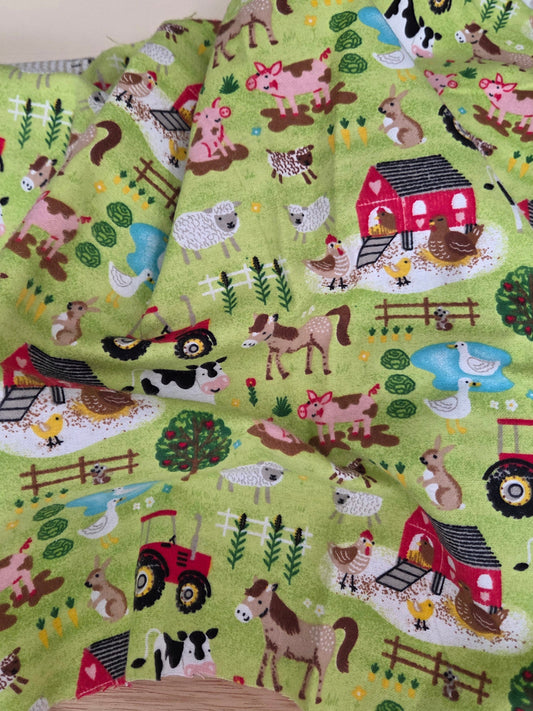 A folded piece of light green fabric with a colorful farm animal and vehicle print, displayed on a wooden surface next to a wooden ruler. LA FInch Fabrics