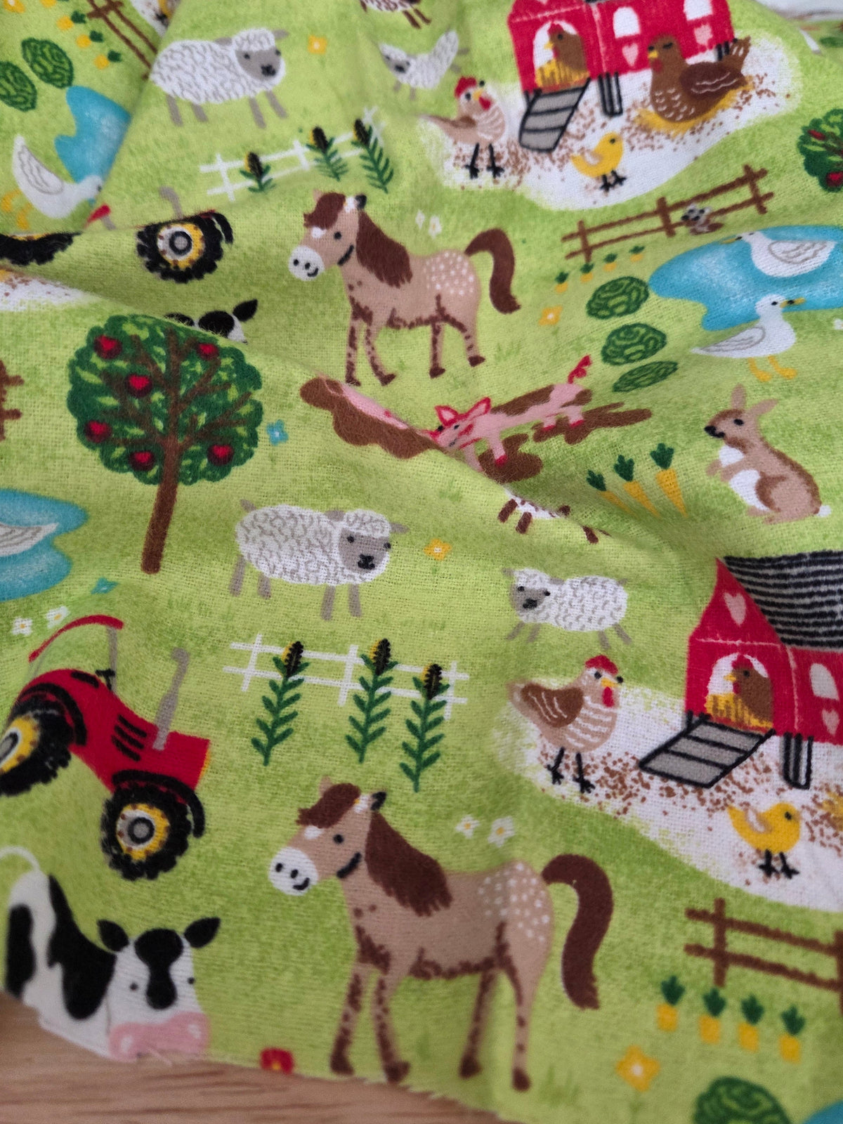A close-up of a fabric product featuring a colorful cartoon farm print on a light green background, showing animals, barns, trees, fences, and vehicles. LA Finch Fabrics