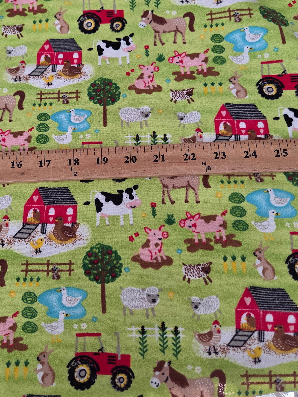 A close-up of a light green fabric with a colorful cartoon farm print, featuring red tractors, cows, horses, pigs, sheep, ducks, and barns. A wooden ruler is placed horizontally across the fabric to show the pattern scale.