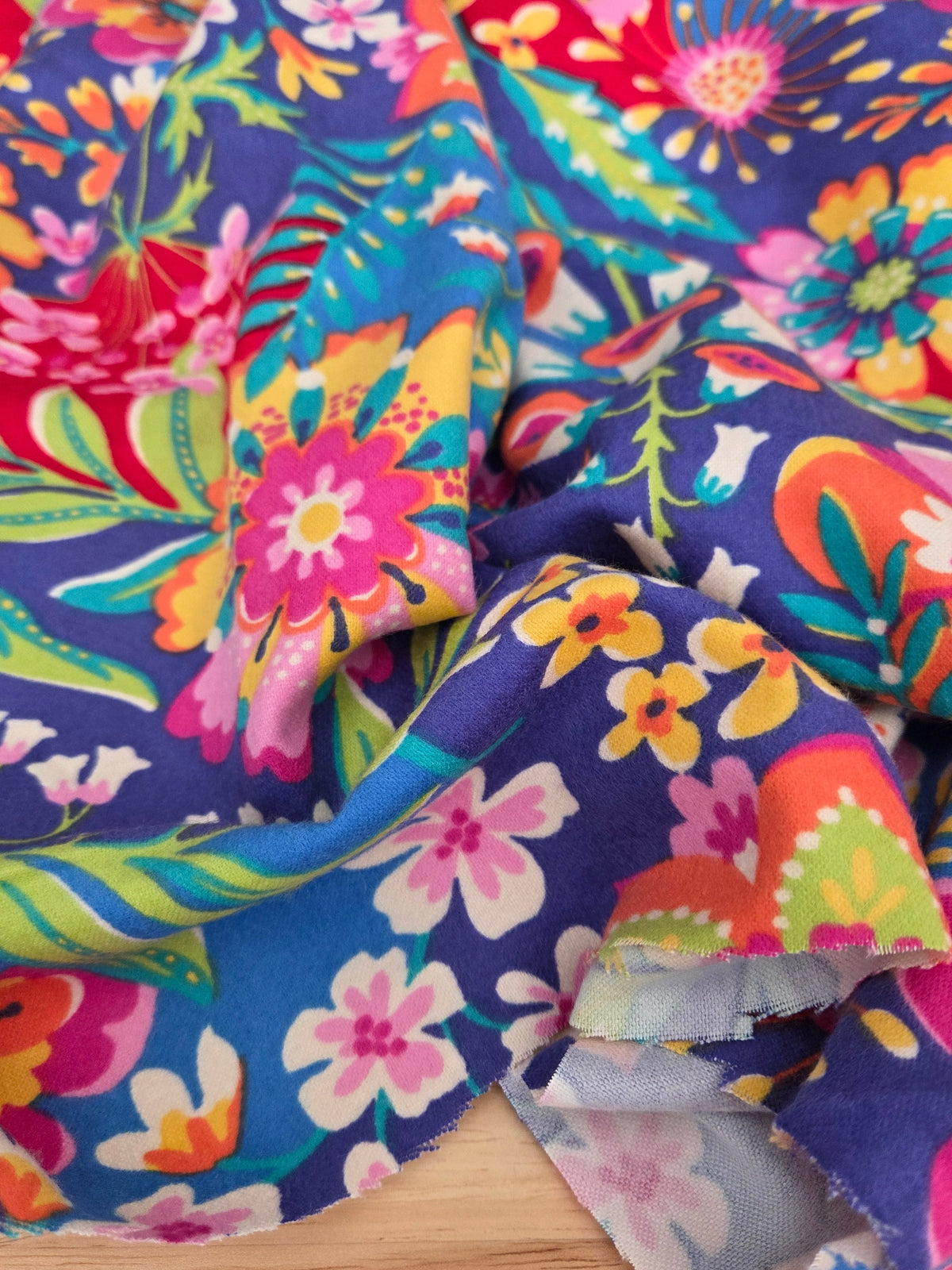 A close-up of a vibrant floral fabric with a multicolor print of pink, yellow, orange, and green flowers on a deep blue-purple background, draped over a light wood surface.