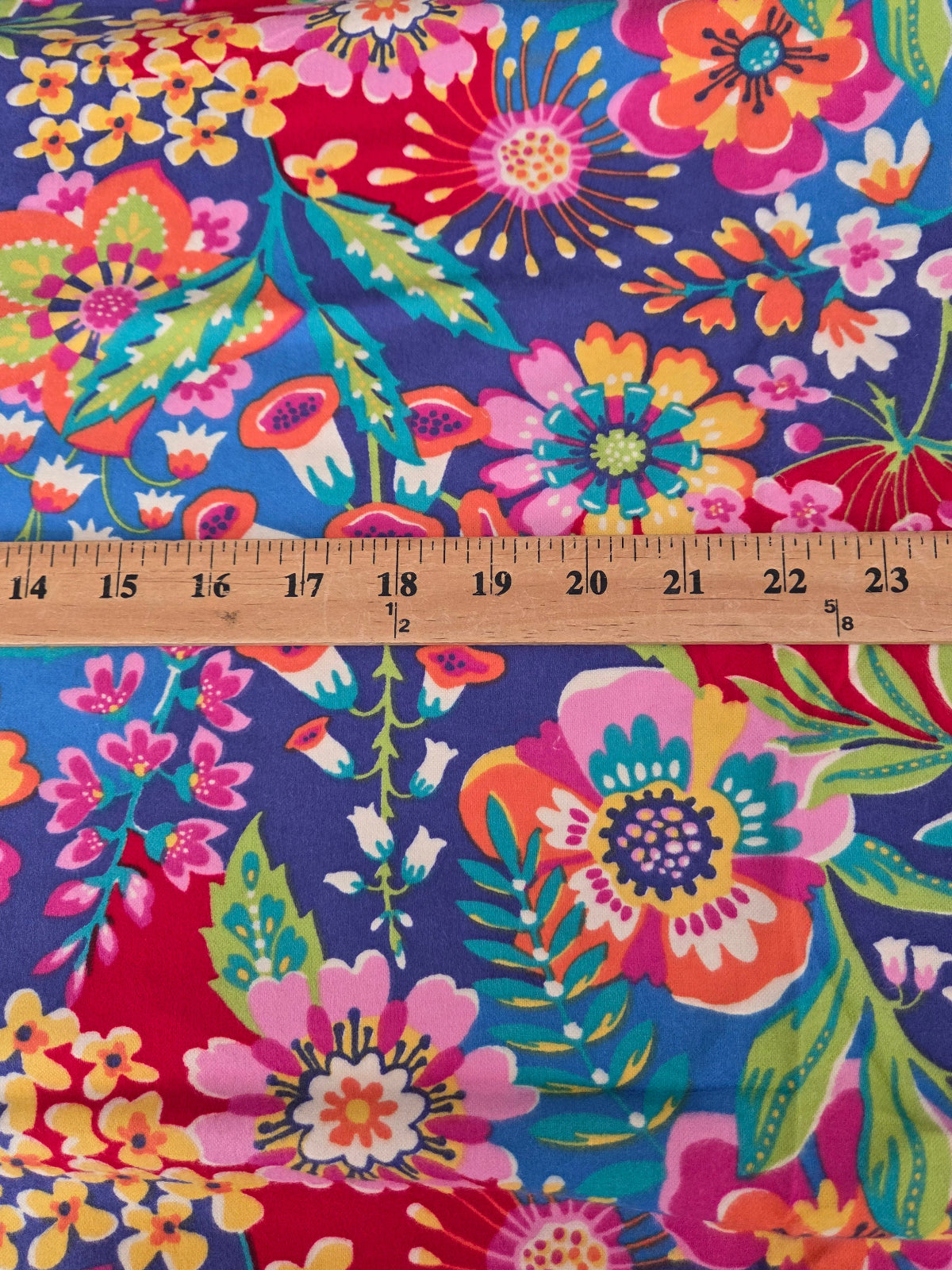 A close-up of a colorful floral fabric with a wooden ruler laid horizontally across the middle to show the pattern scale. The fabric features a dense, vibrant floral print with pink, orange, yellow, green, and white flowers on a deep blue-purple background.