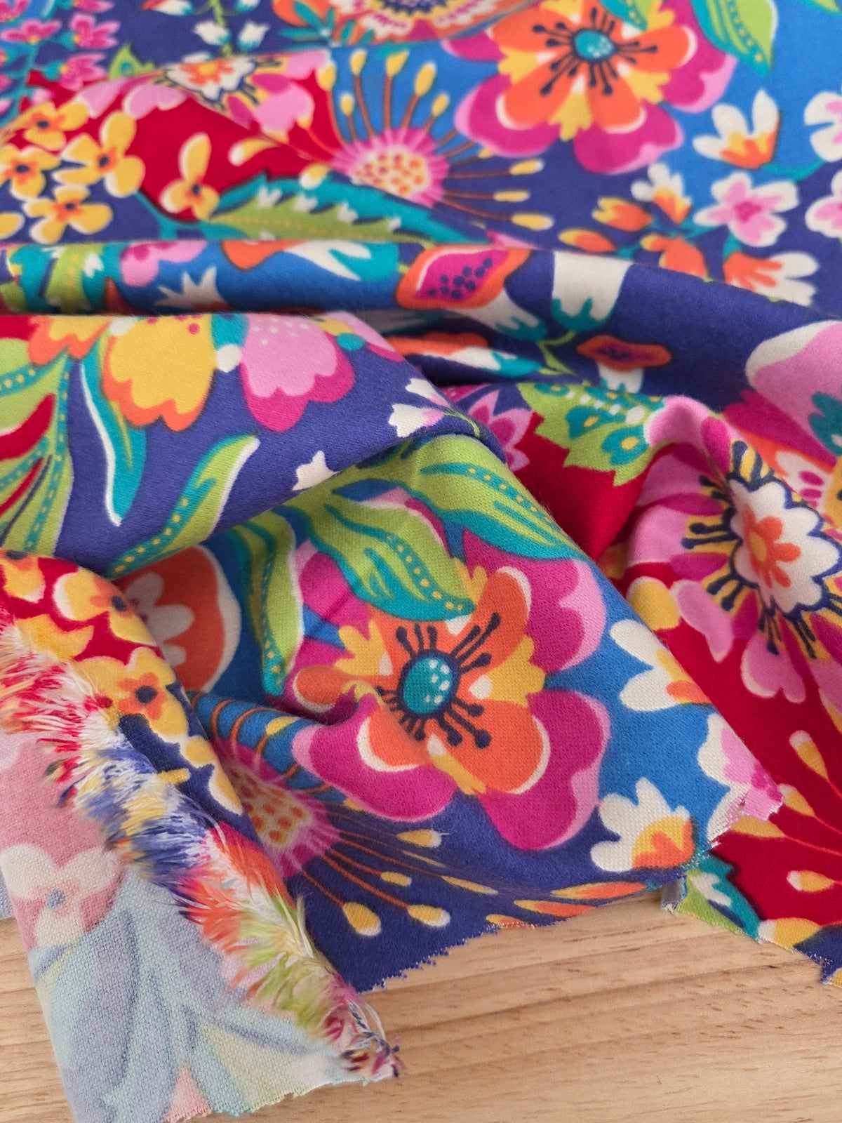 A close-up of a vibrant floral fabric with a multicolor print of pink, orange, yellow, green, and white flowers on a blue and red background, draped over a light wood surface. LA Finch Fabrics