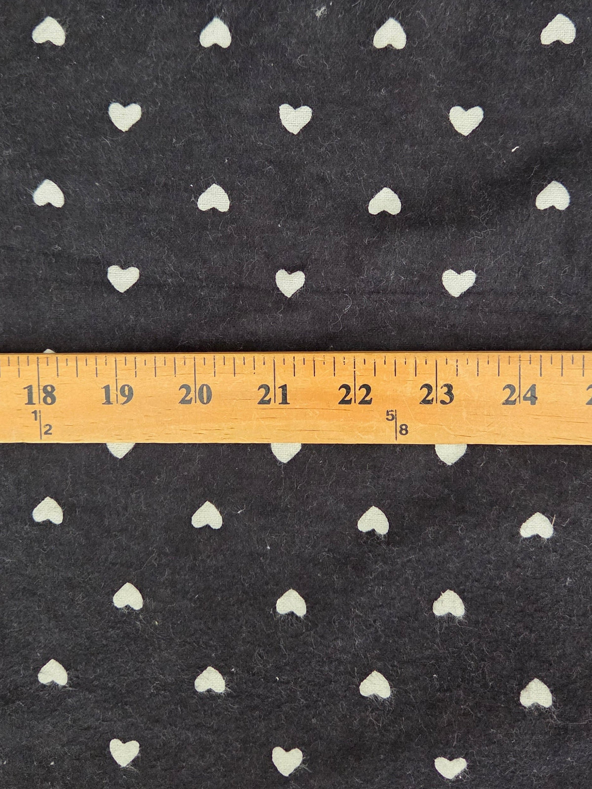 Black fabric with a repeating pattern of small white hearts, shown with a wooden ruler across the middle to indicate scale.