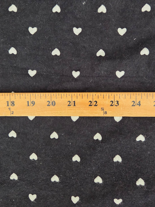 Black fabric with a repeating pattern of small white hearts, shown with a wooden ruler across the middle to indicate scale.