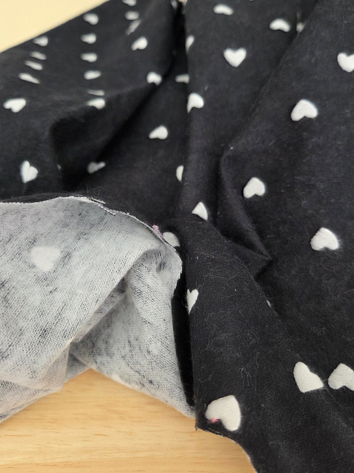 Black fabric with a white heart pattern, folded over to show both the patterned side and a lighter gray reverse side, resting on a light wood surface.