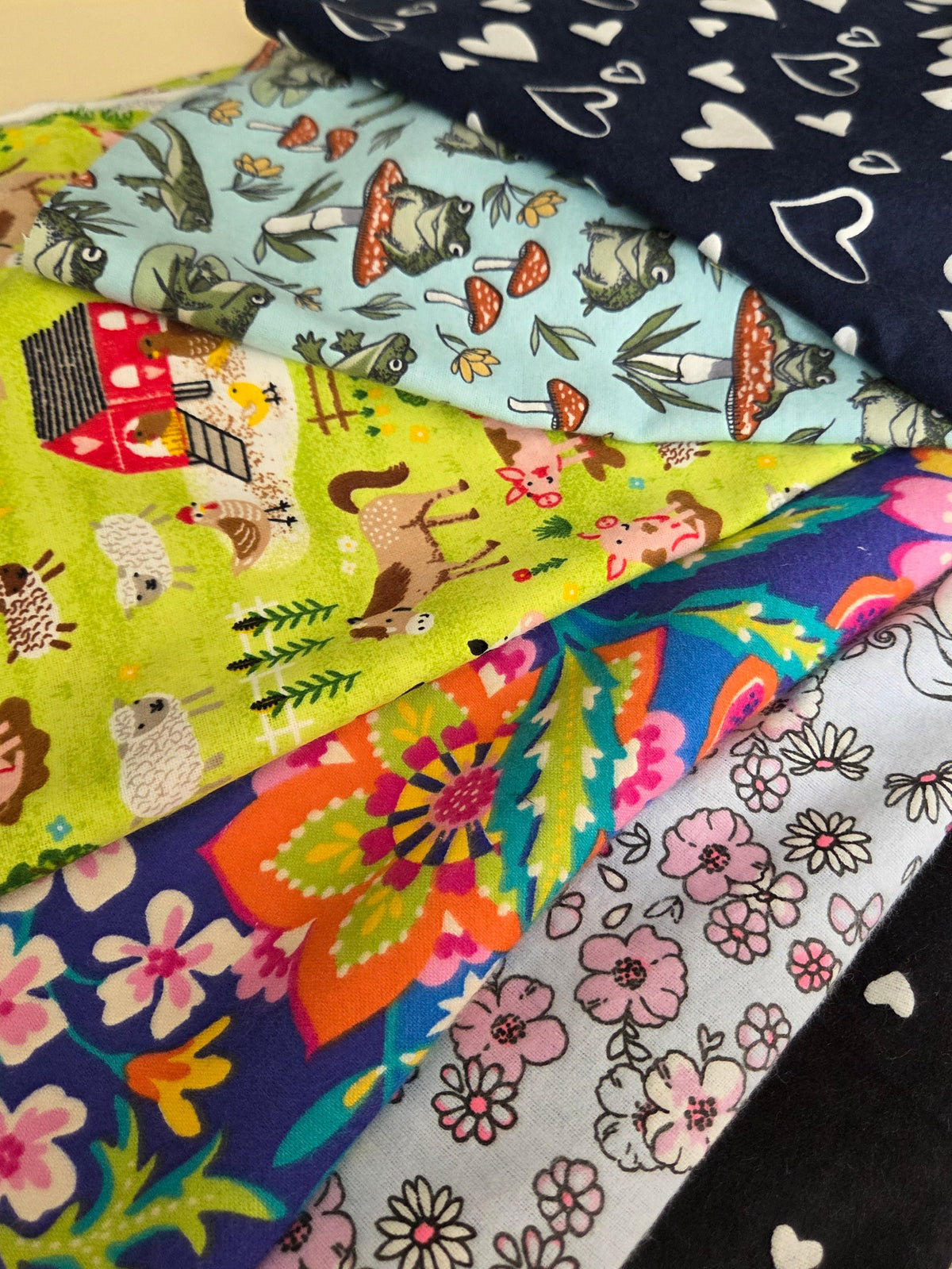 A close-up of several folded patterned fabric pieces layered on top of each other, showing different prints including white hearts on navy, woodland animals on light blue, colorful flowers, and pink floral motifs on black and white fabrics.