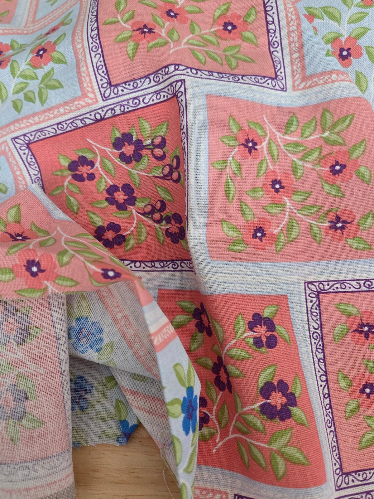 A close-up of a patterned textile laid out on a surface, featuring coral pink squares with green leaves and purple flowers, bordered by pale blue and white sections with ornate detailing.