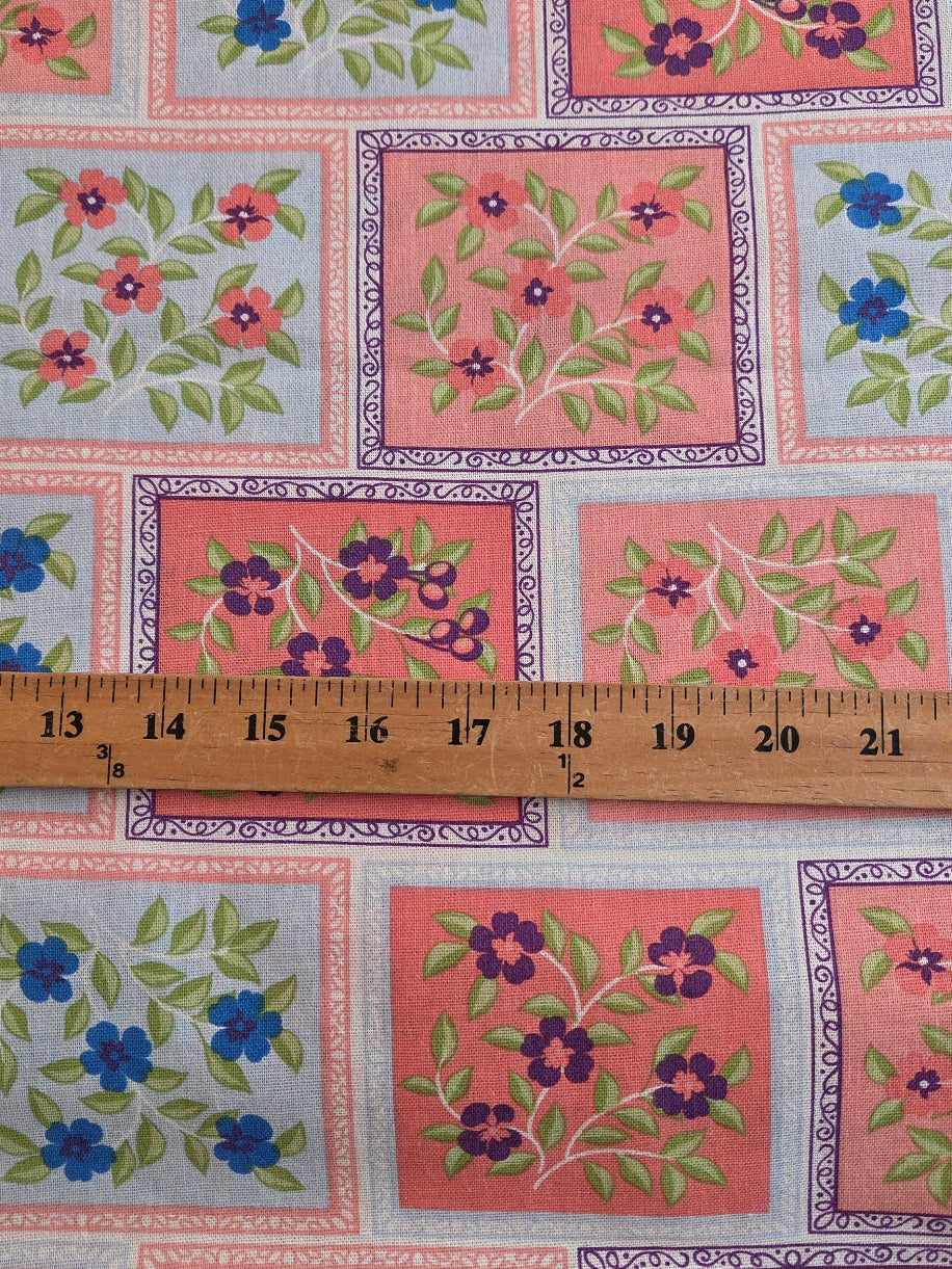 A close-up of a printed fabric with a patchwork-style floral pattern, shown with a wooden ruler across the middle for scale. The design features coral pink, light blue, and white squares with green leaves and purple and blue flowers.