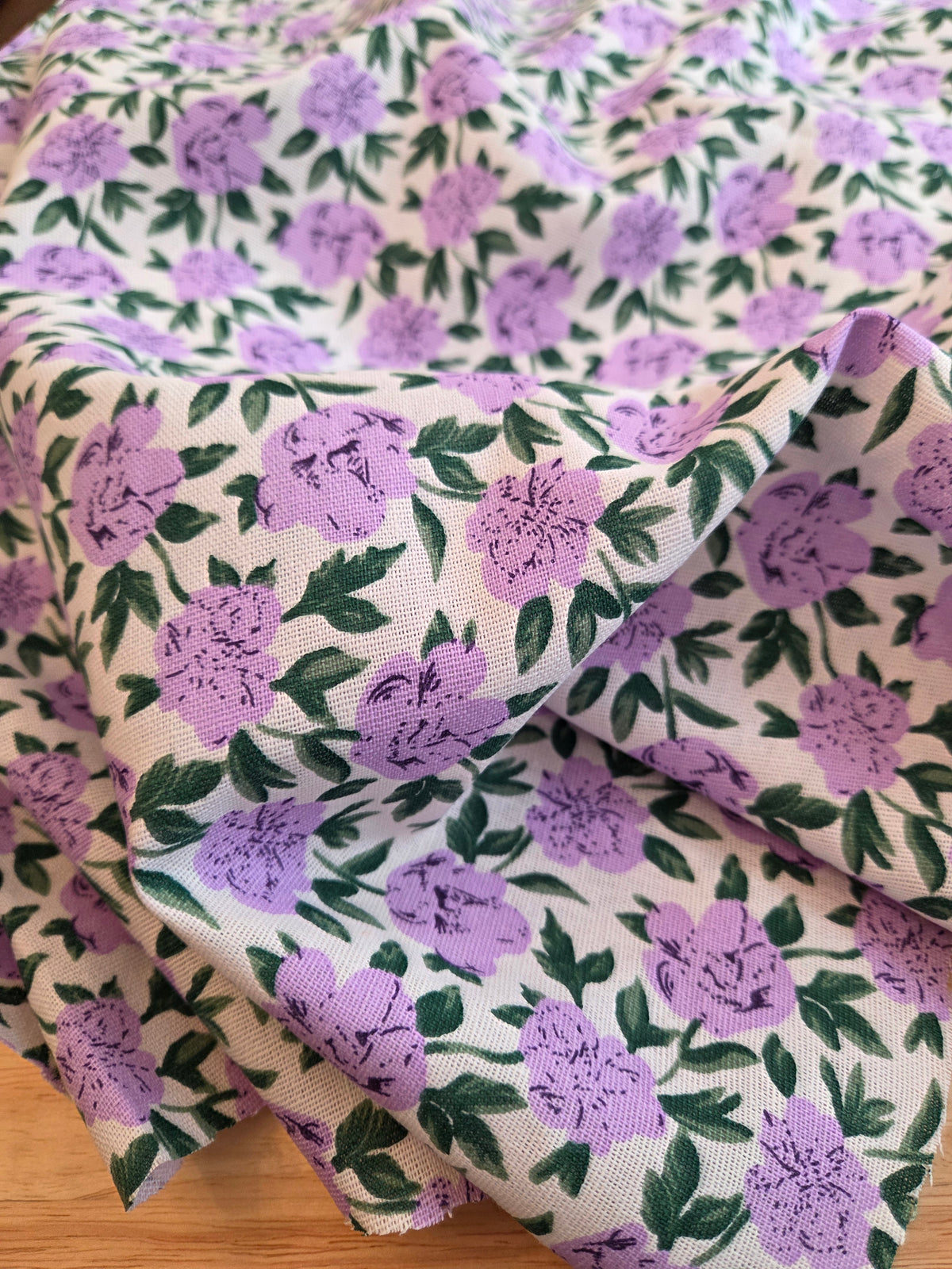 A close-up of draped fabric with a repeating floral print featuring purple flowers and green leaves on a warm cream background.