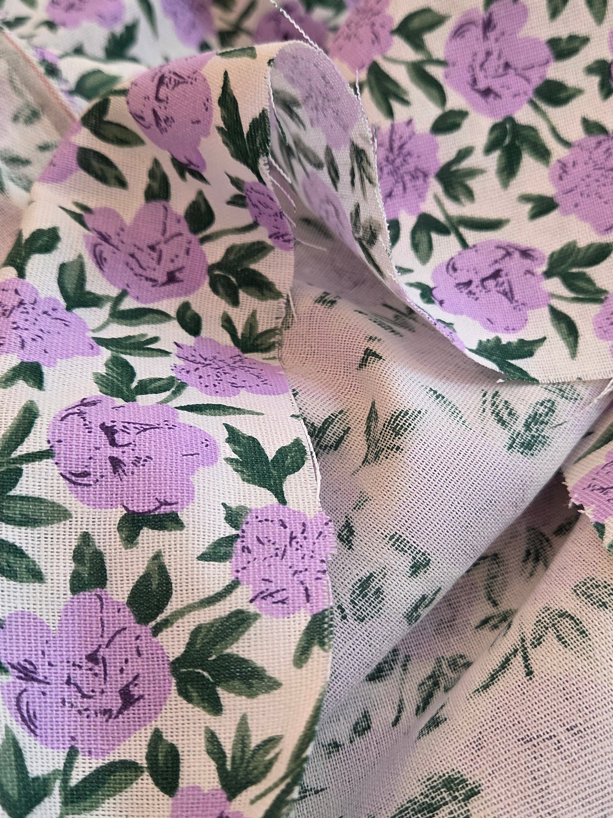 A close-up of patterned fabric with a cream background, purple floral print, and green leaves, with one corner folded over to show the texture.