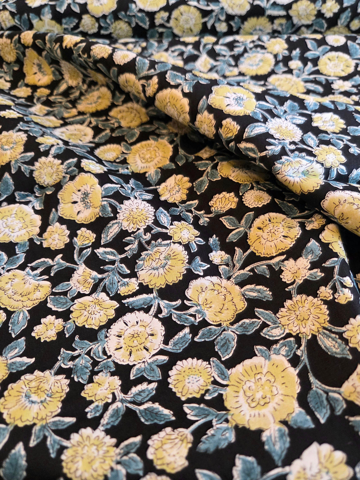 A close-up of draped fabric with a dense floral print featuring yellow and cream flowers with blue-gray leaves on a black background. LA FInch Fabrics