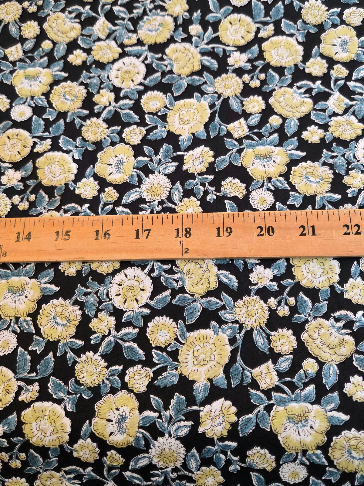 Black floral fabric with yellow and white flowers and blue-gray leaves, shown with a wooden ruler across the middle to indicate scale. LA Finch Fabrics
