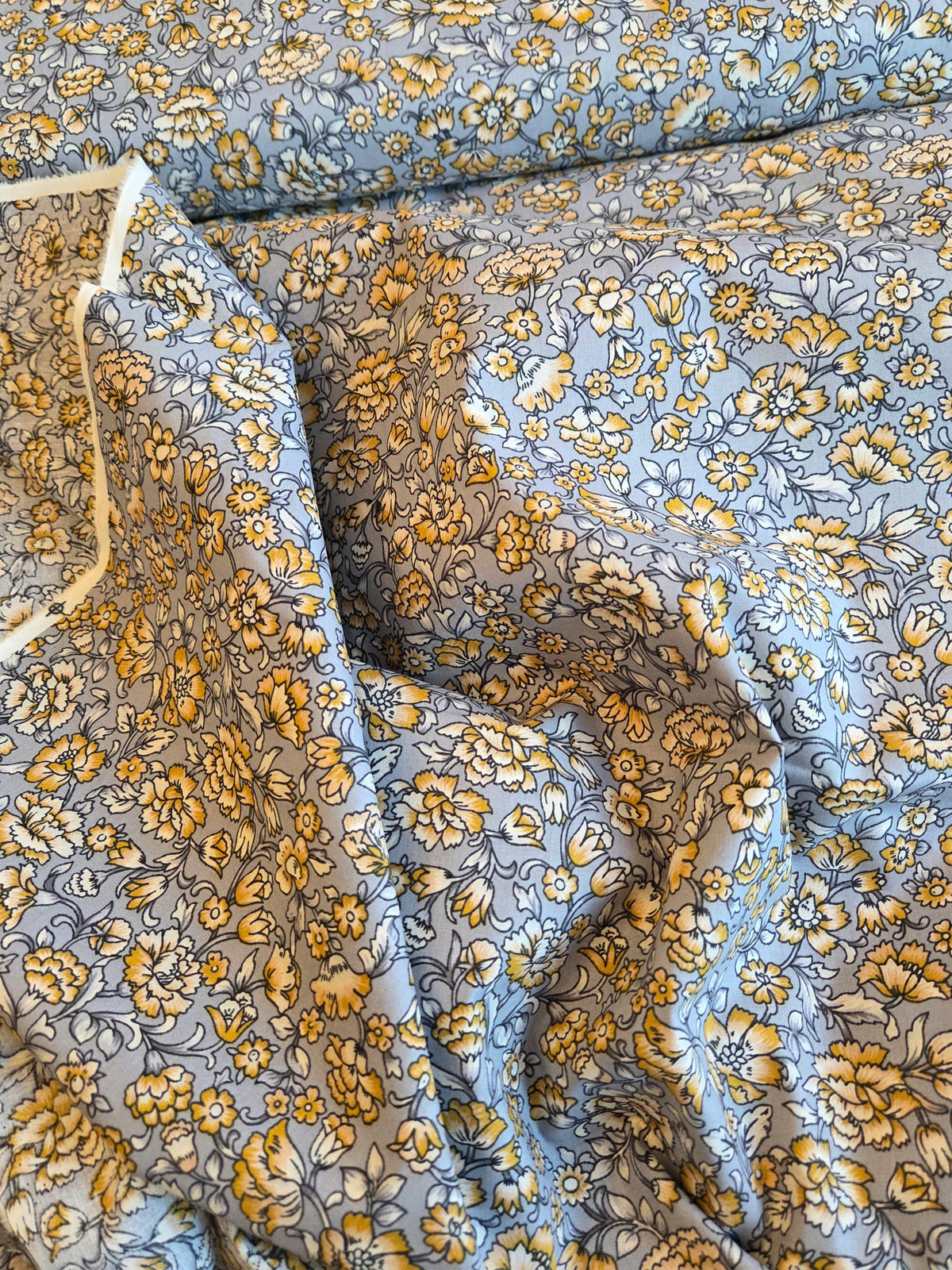 A close-up of a light blue fabric with a dense yellow and white floral pattern, draped with a white plastic hanger visible on the left side. LA Finch Fabrics