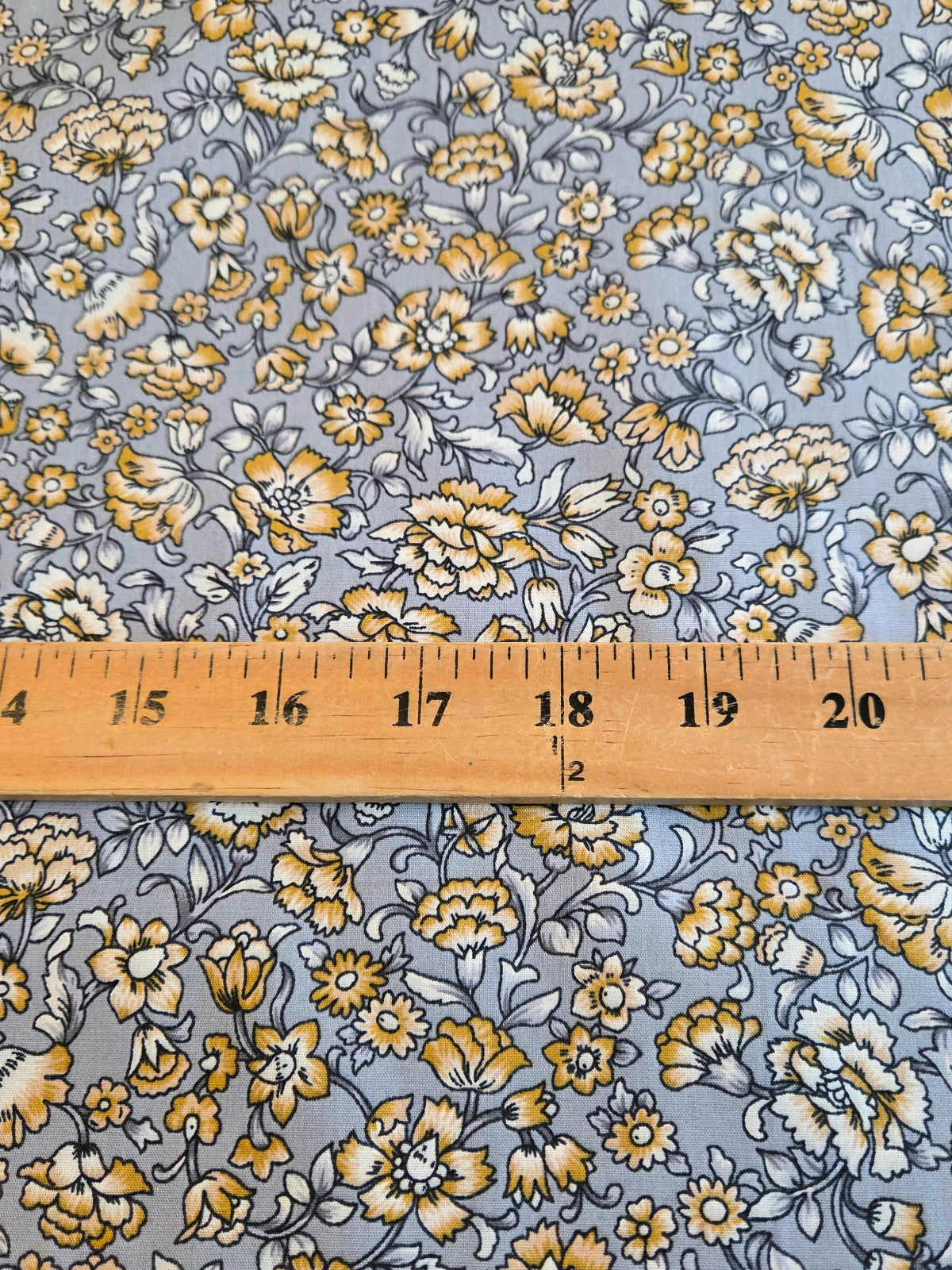 A close-up of floral fabric with a repeating print of yellow, white, and gray flowers on a muted blue-gray background, with a wooden ruler laid horizontally across the middle for scale. LA Finch Fabrics