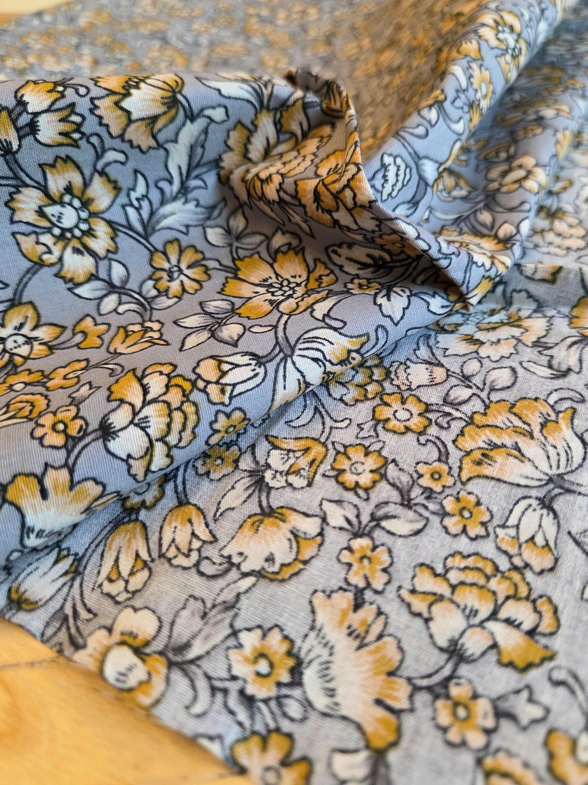 A close-up of draped fabric with a floral print in muted blue-gray, mustard yellow, cream, and dark gray outlines, resting on a light wood surface.