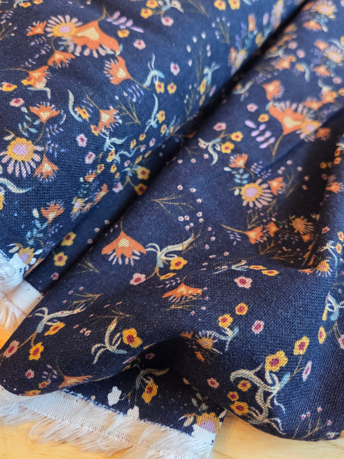 A close-up of dark navy blue fabric with a dense, multicolored floral and botanical print, featuring white fringe along the edge, laid on a light wood surface.