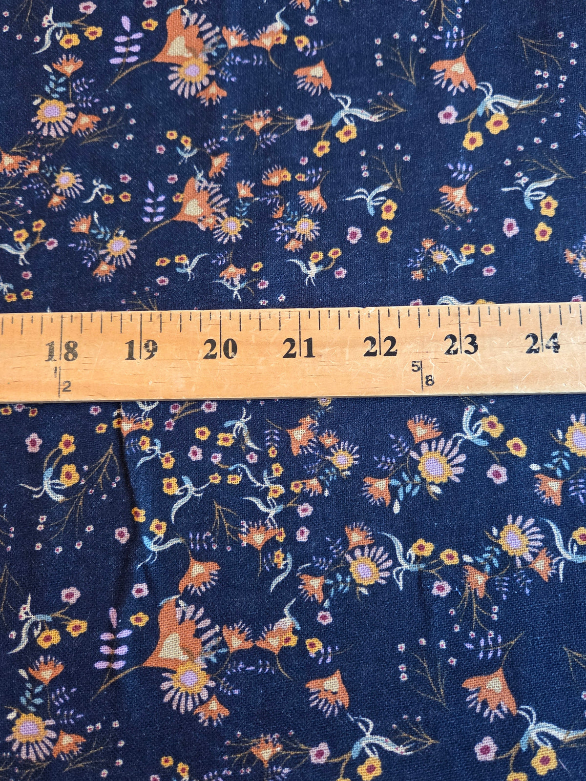 A close-up of navy blue fabric with a dense floral and bird print in orange, yellow, white, and light blue, with a wooden ruler laid horizontally across the center for scale. LA Finch Fabrics