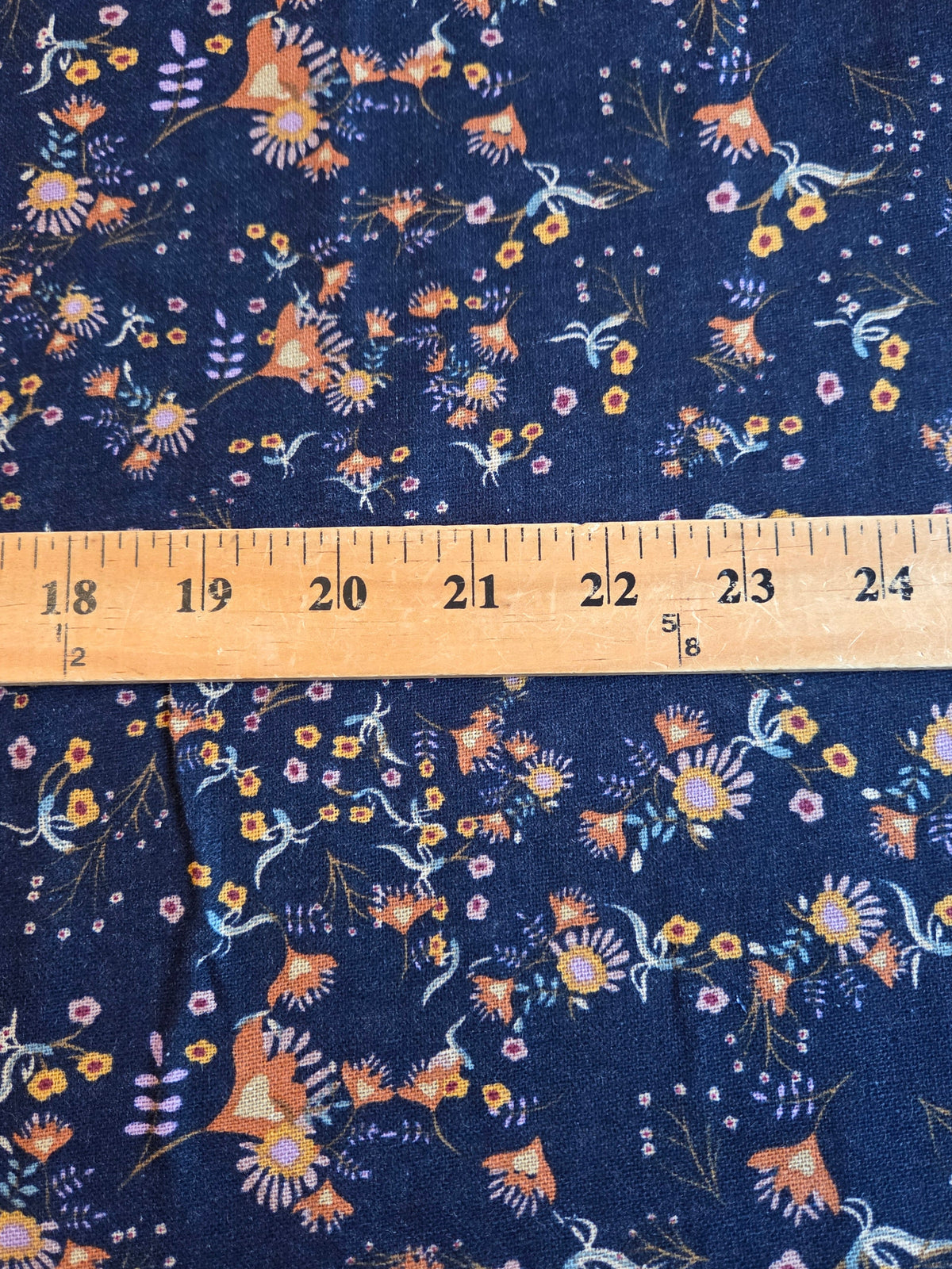 A close-up of navy blue fabric with a dense floral and bird print in orange, yellow, white, and light blue, with a wooden ruler laid horizontally across the center for scale.