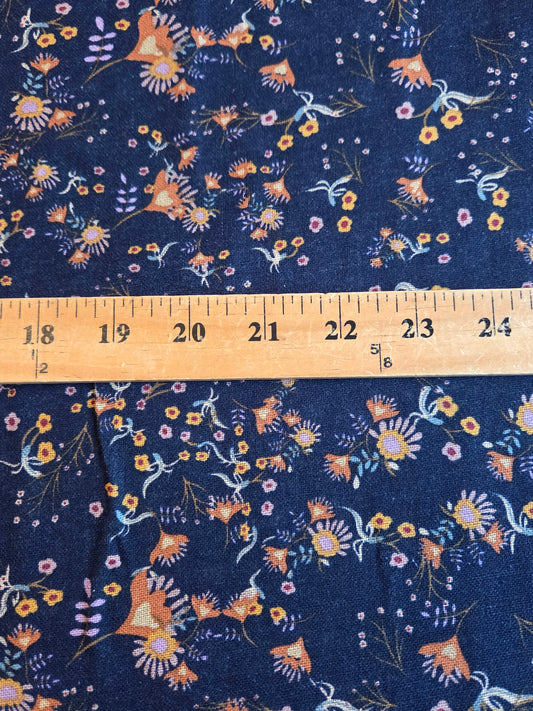A close-up of navy blue fabric with a dense floral and bird print in orange, yellow, white, and light blue, with a wooden ruler laid horizontally across the center for scale.