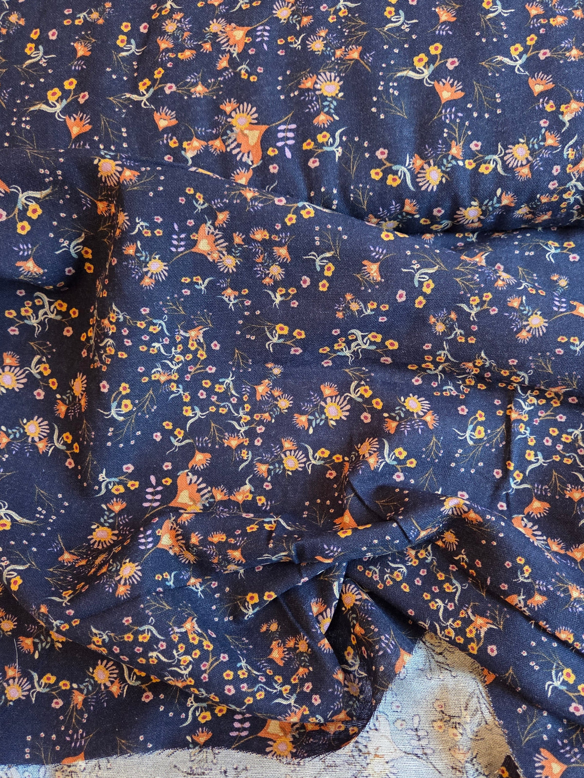 A close-up of a navy blue fabric with an all-over small floral and botanical print in orange, yellow, and light blue, with a second light gray patterned fabric partially visible underneath.