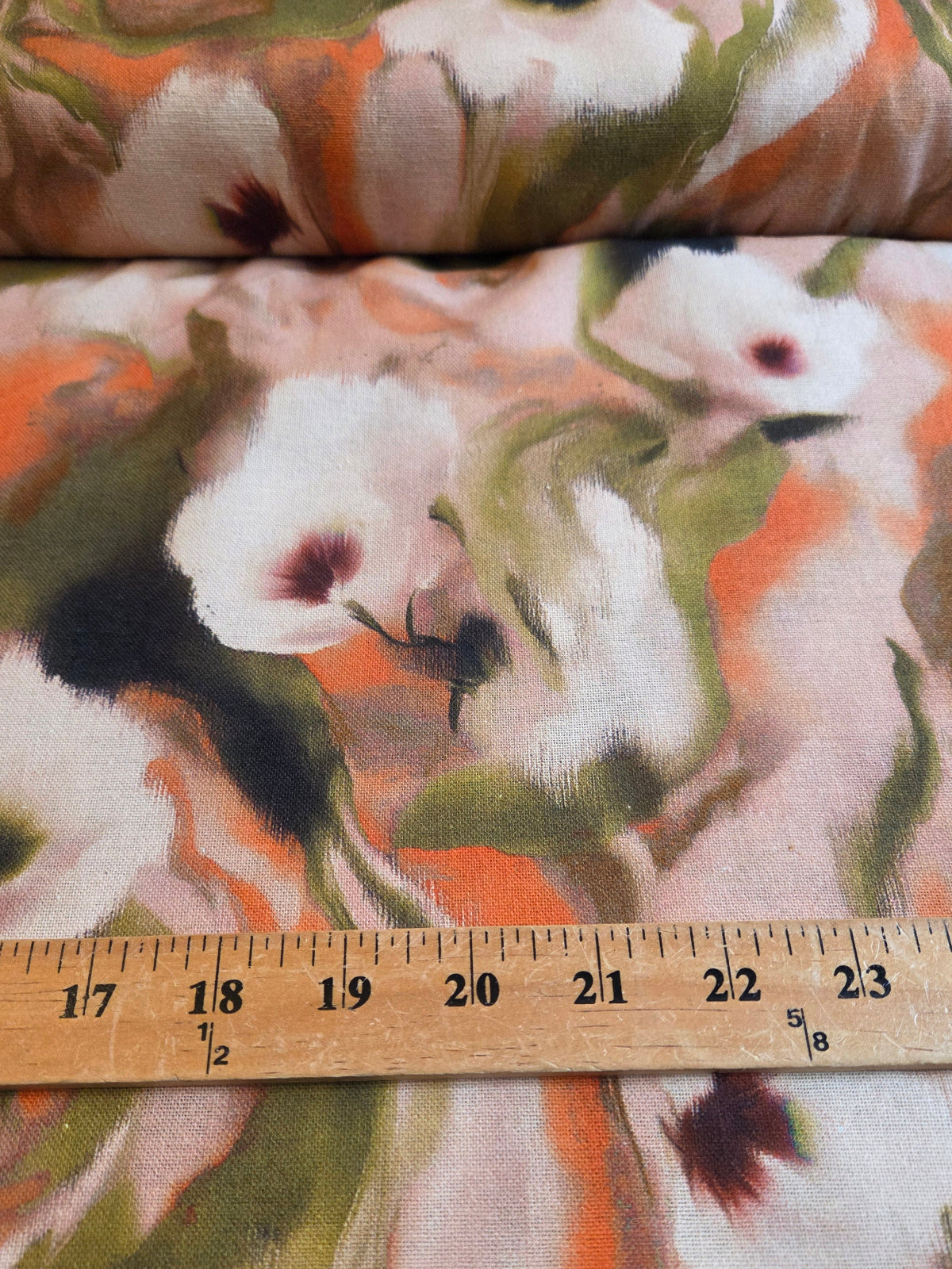 A close-up of floral fabric with large white flowers, orange and green painterly leaves, and a wooden ruler laid across the bottom for scale.