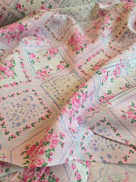A close-up of a draped fabric with a vintage patchwork design featuring pink roses, green leaves, and blue floral motifs on a light cream background. LA Finch Fabrics