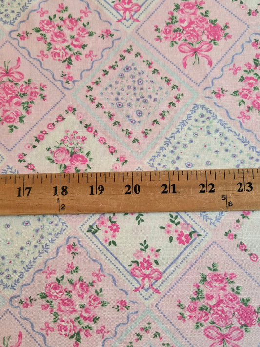 A close-up of a vintage-style floral fabric with a patchwork diamond pattern in pink, blue, and cream, featuring a wooden ruler laid horizontally to show scale.