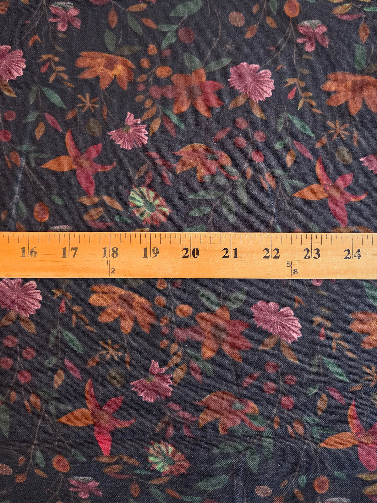 A close-up of floral fabric with a repeating autumn pattern on a dark background, shown with a wooden ruler across the middle for scale.