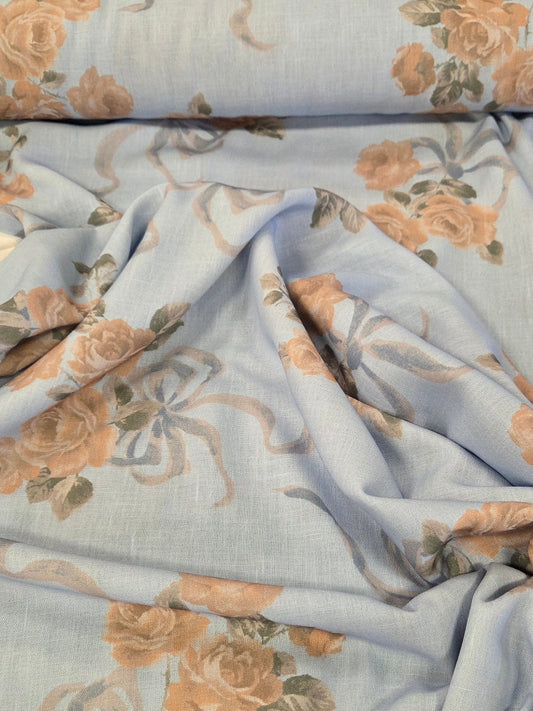 Light blue fabric with a floral print of peach roses and gray bows, draped and folded on a green cutting mat. LA Finch Fabrics
