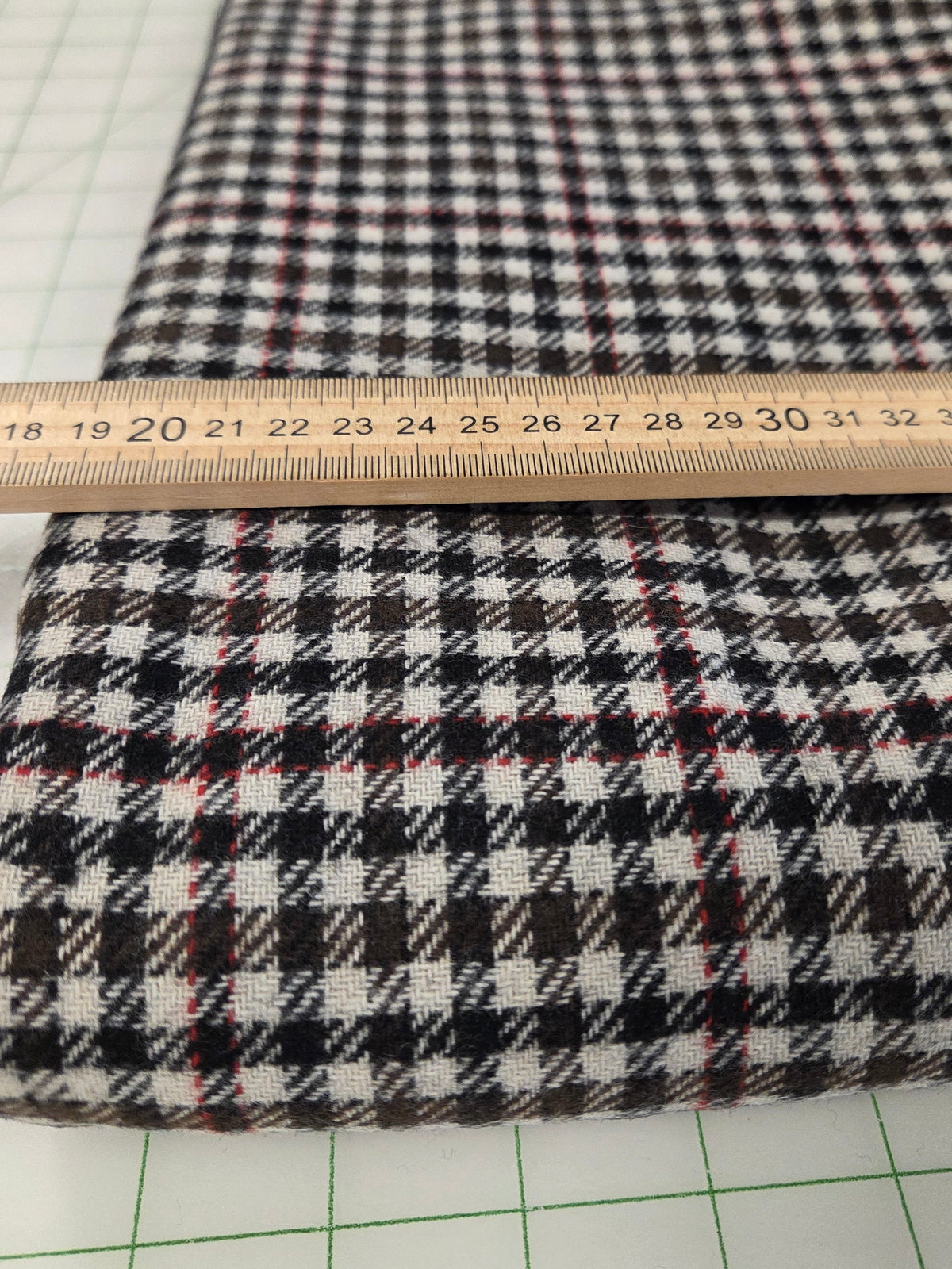 A folded plaid fabric with a black, white, and brown houndstooth pattern and thin red accent lines, shown with a wooden ruler to indicate scale on a green grid cutting mat.