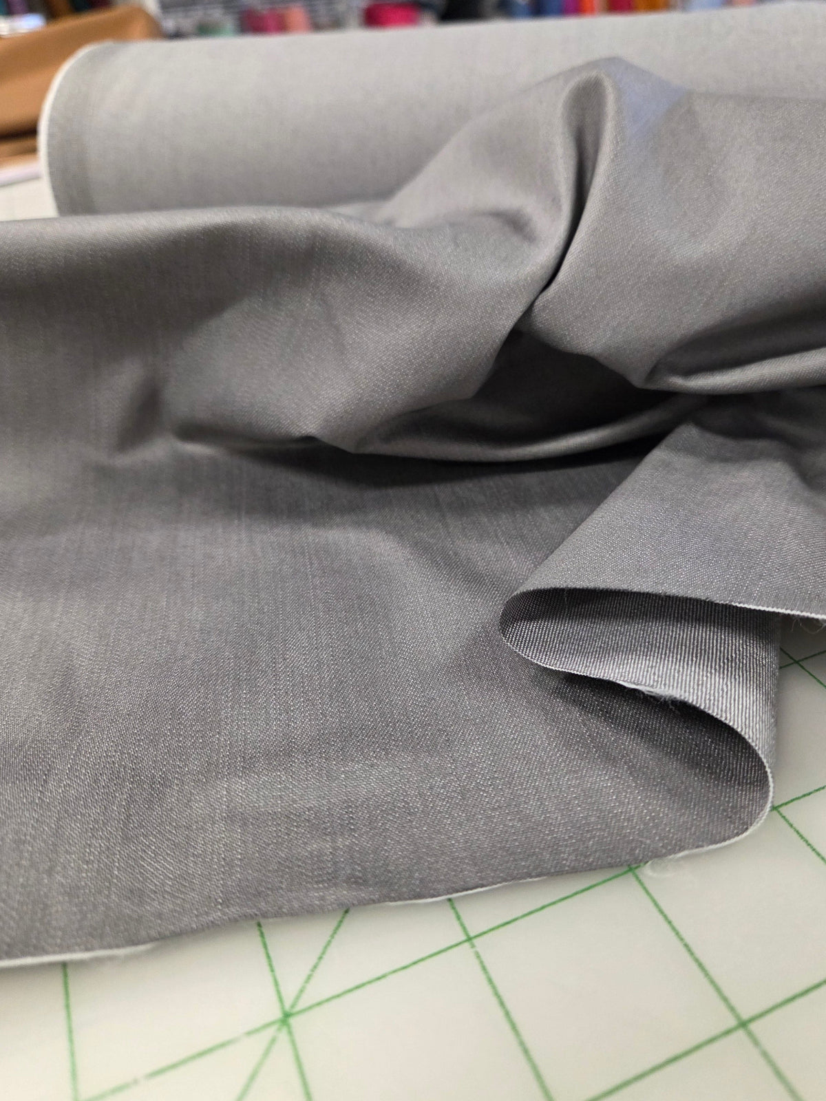 A close-up of draped gray fabric spread across a white cutting mat with green grid lines, with a roll of matching fabric in the background and a blurred craft-supplies environment behind it. Japan Fabric light gray for garment making sewing