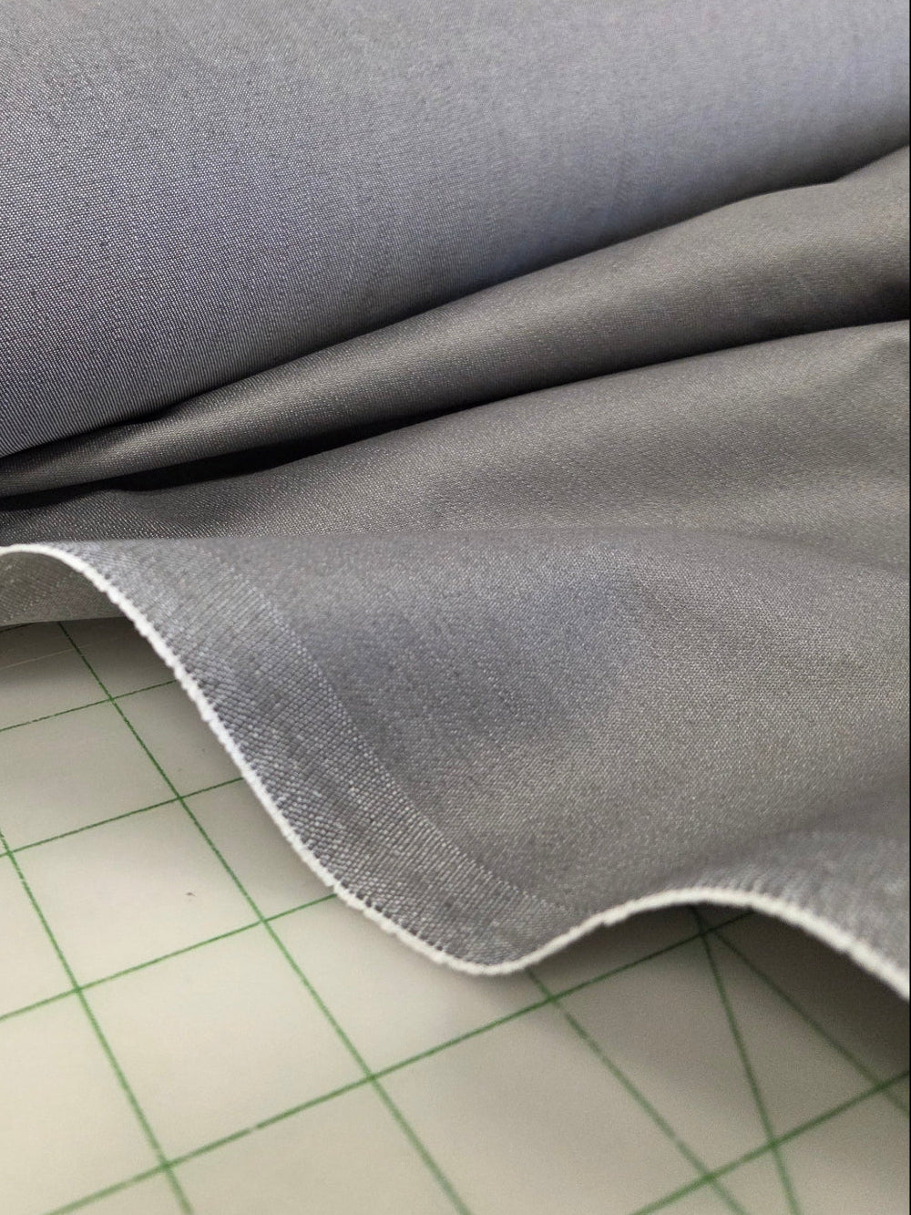 A close-up of a light gray woven fabric roll draped over a white gridded cutting mat, with a fabric store or studio interior visible in the blurred background.