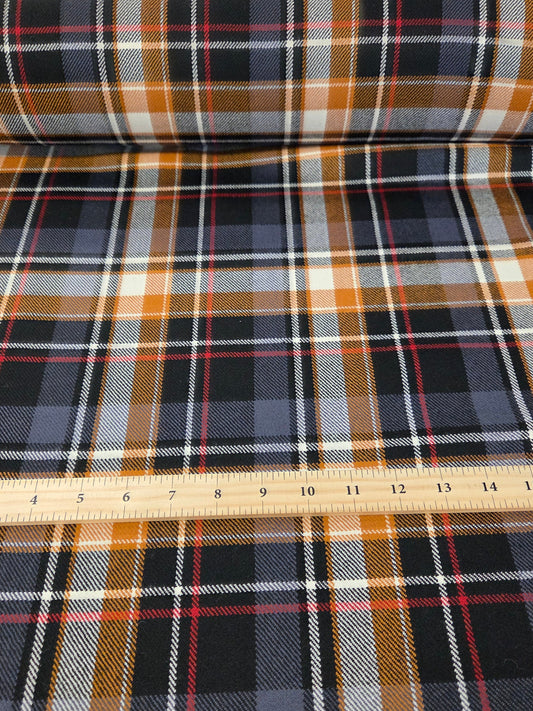 A roll of plaid fabric shown flat, with a wooden ruler laid across the center to indicate scale. The fabric pattern features black, navy, gray, white, and rust-orange plaid stripes. wool suiting blend for garment making. fabric roll
