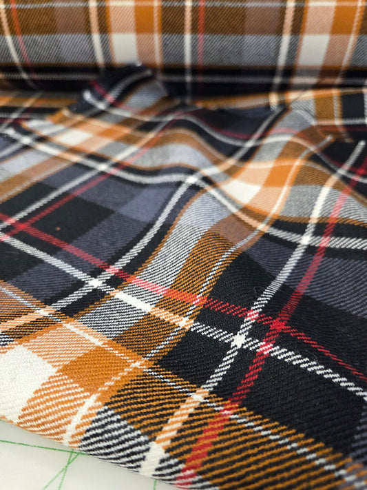 A close-up of folded plaid fabric with a tartan pattern in black, orange, white, gray, and thin red lines, resting on a white cutting mat with green grid markings. wool suiting