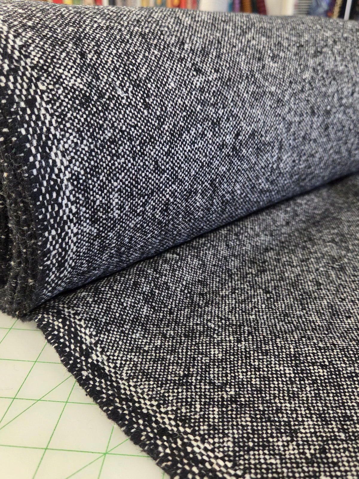 A close-up of a dark charcoal gray and white heathered woven fabric, shown partially rolled over a green grid cutting mat with a fabric store background visible. wool tweed woven for coats