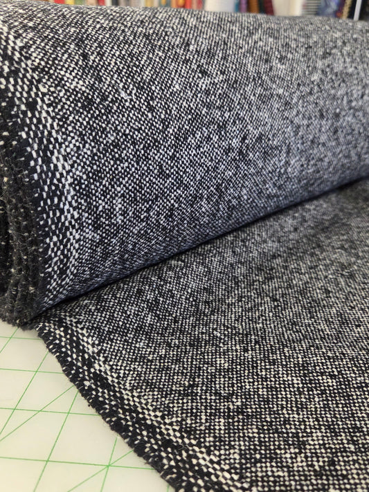 A close-up of a dark charcoal gray and white heathered woven fabric, shown partially rolled over a green grid cutting mat with a fabric store background visible. wool tweed woven for coats