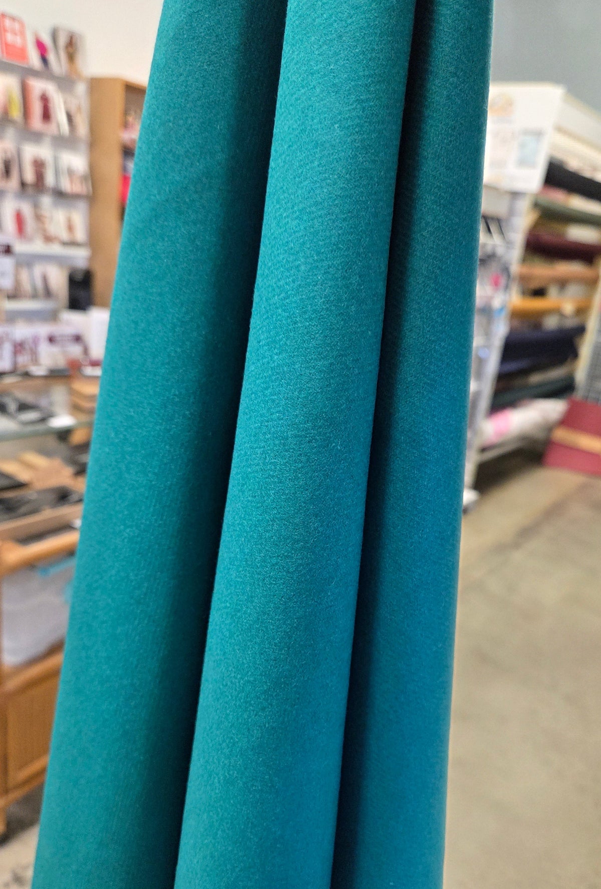 A close-up of teal blue fabric hanging vertically in folds, with a blurred store interior visible in the background. wool italian fabric coating
