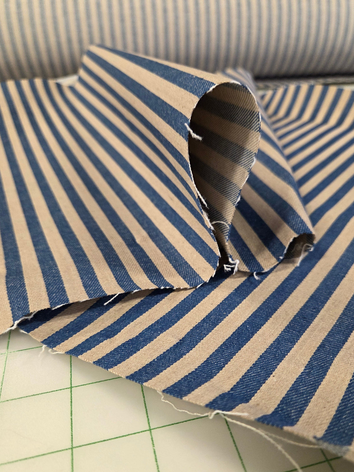 Blue and beige striped fabric folded on a green grid cutting mat, with a matching striped fabric roll visible in the background. Denim Cone Mills Fabric Stripe for garment making sewing. Price by the yard