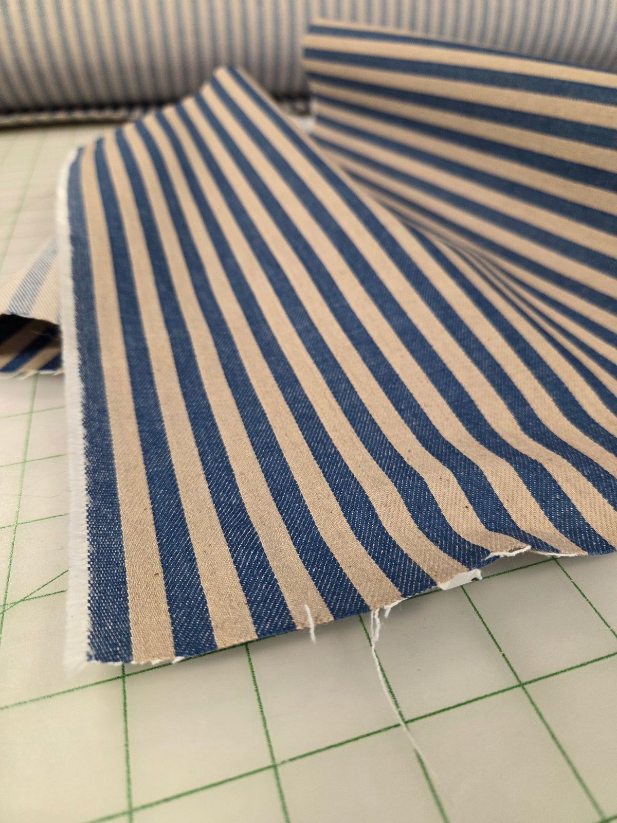 Blue and beige striped fabric with raw, frayed edges, laid on a green-lined cutting mat. Cone Mills Denim Deadstock Mill Fabric by the yard. 