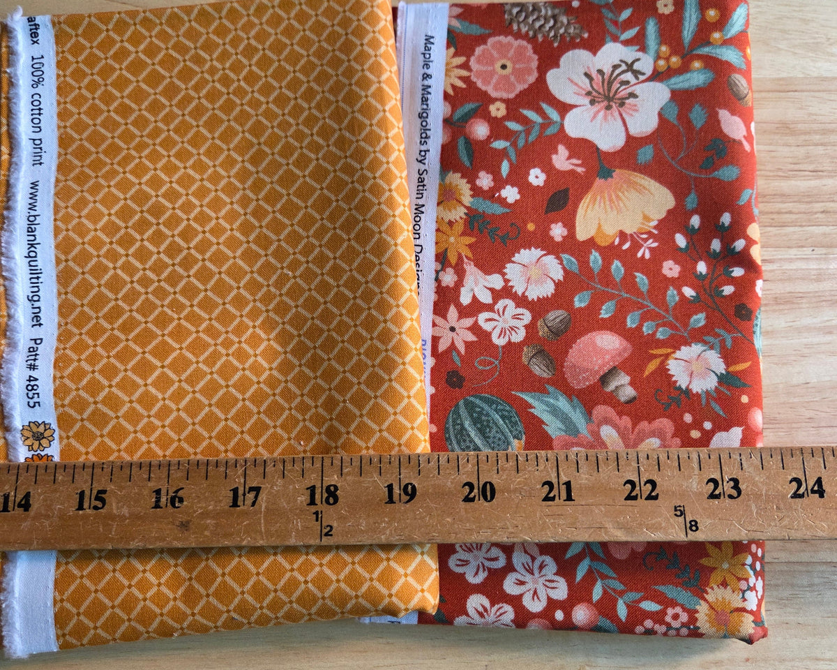 Two folded pieces of fabric laid on a wooden board, with a wooden ruler placed across them to show size. The left fabric is mustard yellow with a white geometric trellis pattern.